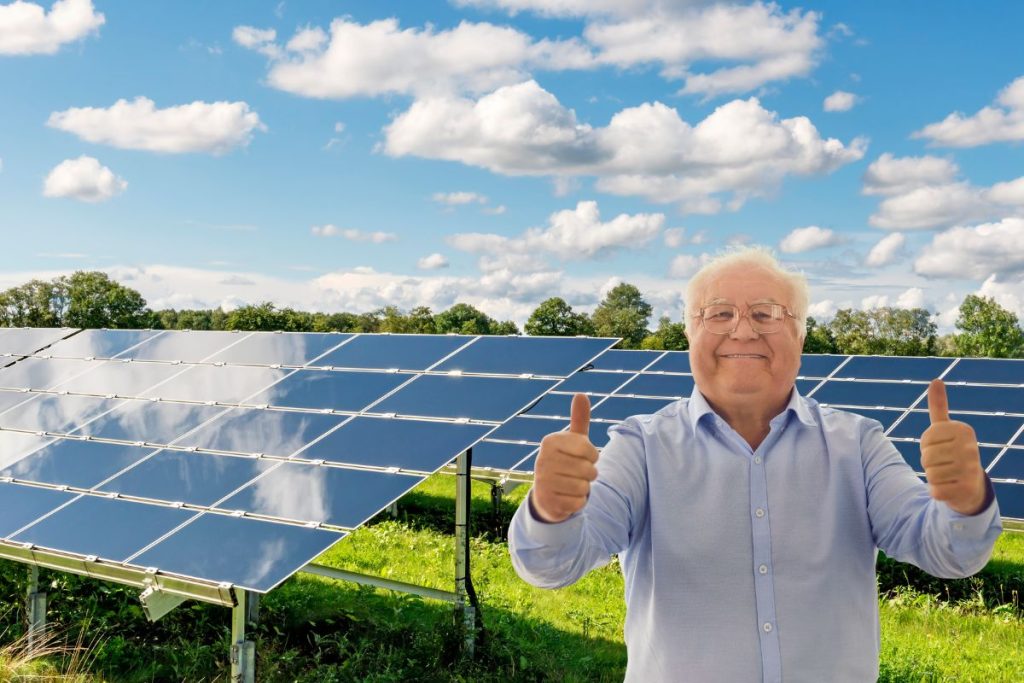 Zadovoljan stariji muškarac pokazuje palčeve gore ispred solarnih panela montiranih na zemlji, na sunčanom danu u prirodnom okruženju.
